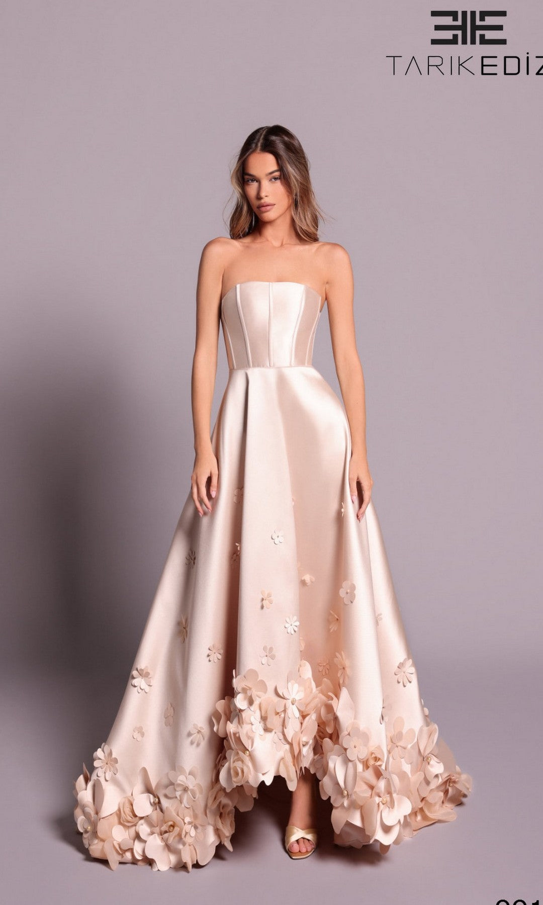 A woman wears the Tarik Ediz 99146 long formal dress in champagne, featuring a strapless fitted bodice and 3D floral embellishments along the hem for an elegant touch.